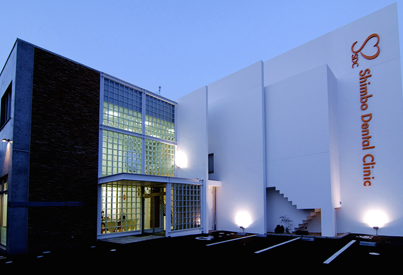 夕暮れ時の医院の外観写真。地面からのライトアップで、白い壁にはオレンジ色の「しんぼ歯科クリニック」の文字が浮かび上がるように照らされています。