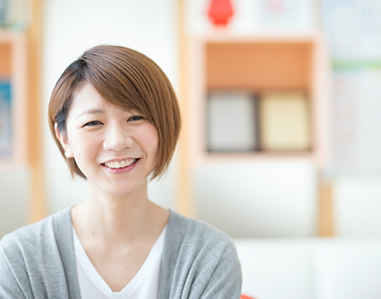 白いシャツとグレーのカーディガンを着た女性の患者さんがカメラに笑顔を向けている写真。背景は明るくぼやけており、院内の様子が写っています。